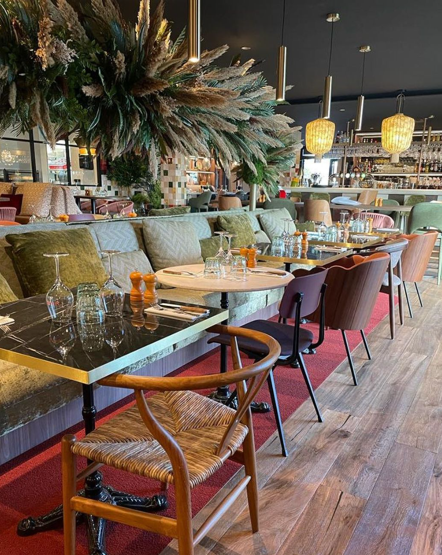 Salle de restaurant chic avec tables en marbre, chaises variées, banquettes vertes, tapis rouge et décoration végétale suspendue. Ambiance moderne et raffinée.